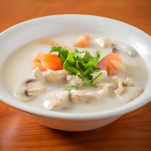 Close-up of a creamy Thai soup with chicken, mushrooms, tomatoes, and cilantro in a white bowl.