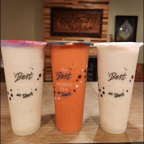 Three SaiMai Thai Restaurant bubble tea drinks, two light-colored and one orange, on a wooden surface.